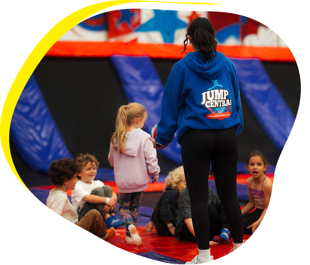 Adult staff member in a blue “Jump Central” hoodie holding a young child’s hand while several children sit and play on padded mats inside an indoor trampoline park.