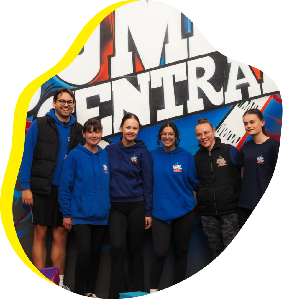 Group of staff members wearing blue hoodies standing together and smiling in front of a colorful “Jump Central” mural wall.