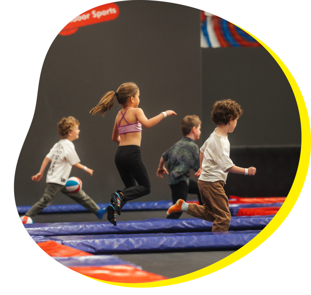 Group of young children running and jumping across padded trampolines at an indoor trampoline park, with one child holding a small red and blue ball.